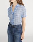 A woman models the FRAME Essential Top—a lightweight blue plaid button-front style—tucked into high-waisted blue jeans, standing against a plain light background with her face partially out of frame.