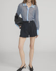 A woman wears The Collared Cardi by FRAME over a sheer blue and white striped shirt with high-waisted black shorts, black slingback heels, and a black clutch, styled against a plain background.