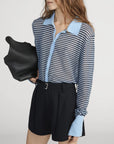 A woman with wavy brown hair wears FRAME’s The Collared Cardi, a sheer blue and navy striped cardigan with a light blue collar and cuffs, paired with high-waisted black shorts and carrying a large black bag.