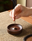 A hand places a lit incense stick into the Faire Walnut Wood Incense Burner on a woven placemat, with smoke rising gentlyโshowcasing traditional Chinese artistry and a unique rustic glaze.