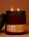 The Faire Howdy Honey Candle is a brown coconut soy wax jar candle with two flames, shown beside its lid on a warm brown background.