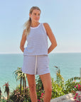 A woman stands outdoors near blooming plants and the ocean, wearing Faire’s Stripe Track Short with a sleeveless blue-and-white striped top.