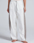 A person stands barefoot in ASKK's Cancun Pant, featuring a wide-leg silhouette, white fabric with gray vertical side panels, and an elastic drawstring waistband, photographed against a plain light background.