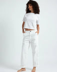 A woman with curly hair wears a white t-shirt and ASKK chino twill relaxed, loose fit pants, standing barefoot against a plain light background with her hands in her pockets, looking slightly to the side.