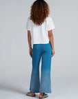 A person with curly hair stands facing away in a loose white short-sleeve top and ASKK Indigo Dip Dye Sailor Sweats, made from American knit cotton in Los Angeles, paired with gray slip-ons. The background is plain light gray.