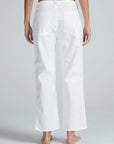 A person stands barefoot, facing away from the camera, wearing ASKK’s Tomboy Twill high-waisted, loose-fit white stretch twill pants with large back pockets and a straight leg against a plain light gray background.