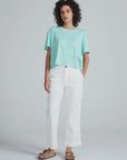 A person with curly hair wears a loose light blue-green t-shirt, ASKK's Tomboy Twill high waist white stretch pants, and beige sandals, standing against a plain light gray background.