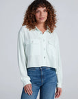 A woman with curly brown hair wears the ASKK Crosby Shirt, featuring light green and white stripes and chest pockets, paired with blue jeans, standing against a plain light background.