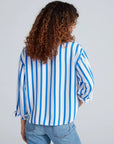 A person with curly brown hair, seen from behind, wears the ASKK Crosby Shirt—a blue and white vertically striped 100% cotton relaxed fit—and light blue jeans against a plain light background.