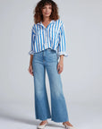 A person wears the ASKK Crosby Shirt, a relaxed fit blue and white vertically striped 100% cotton button-up, paired with light blue flared jeans and white shoes, against a plain background.
