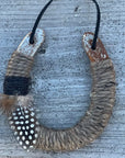 The Lucky Horseshoe by Faire features an upcycled horseshoe wrapped in twine with a black band and spotted feather, hanging from a black cord—a unique housewarming gift displayed against rustic wood.