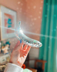 A hand holds up the Faire Disco Banana, a handmade piece covered in tiny mirror tiles that reflect sunlight around a cozy, colorful room with green curtains and framed art.