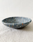 The Kitopo Small Bowl by Faire, a shallow, round Rwandan basket with blue and gray coils plus white and orange accents, rests on textured light gray fabric against a neutral background.
