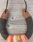 The Lucky Horseshoe by Faire features upcycled horseshoes wrapped in dark gray and multicolored yarn with orange, pink, yellow, and teal accents. Hung by a black cord, this unique piece makes a personalized gift.