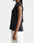 A person stands in profile against a plain background, wearing the Rachel Comey Miles Tee—a sleeveless black top featuring hand crochet stitch detailing on the shoulders—paired with loose white pants and long braided hair for a minimalist look.