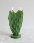 The Nopal Cactus Beeswax Candle by Faire features a green, paddle-shaped cactus design with dual flames, standing upright on a round base—a striking home décor piece shown on a white textured cloth with a plain background.