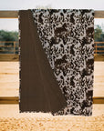 The Faire Bella Hide Brown Quilt, featuring a brown and white cowhide pattern with a solid brown underside, hangs over a wooden fence outdoors, bringing Fort Worth Texas style to any setting.