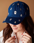 A person with long brown hair smiles slightly, wearing the Faire Dog Days Baseball Cap—a blue cap embroidered with small dog faces and an adjustable strap. The plain background is light brown.