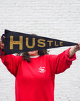 A person in a red sweatshirt holds the Faire Hustle Pennant, showcasing bold gold "HUSTLE" lettering and concealing their face. The backdrop is decorated with white brick wall decor.