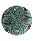 The Birava Large Bowl by Faire is a handmade, round woven tray with a teal spiral pattern, accented with geometric designs in brown, blue, white, and yellow spaced evenly around its edge.