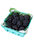 A small turquoise carton of fresh blackberries sits beside Faire’s Blackberry Beeswax Candles, both lined with green and white paper and set against a white background.