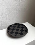 Round glossy black check snack plate by Bungalow Furniture, displayed on a white surface near a textured wall.