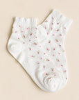 The Faire Blossom Breeze Sheer Floral Ankle Socks are white ankle socks with a subtle ribbed texture and small pink floral designs, shown laid flat on a light beige background.