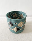 The Blue Storage Plant Basket: Mifuti by Faire features teal, black, and natural geometric patterns. Handwoven in Rwanda, this unique basket planter adds an authentic touch of African decor to any room.