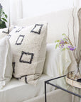 A cozy bed with a POM POM Bowie Big Pillow and neutral bedding sits beside a glass nightstand holding a woven tray, vase with purple flowers, and black decor. A green leafy plant is visible in the background.