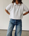 A person wearing Hey Gang’s The Boxy Polo and blue jeans stands hands-on-hips, facing forward against a plain light background, head partially out of frame.