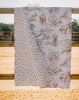 The Faire Buffalo Toile Brown Quilt is draped over a wooden fence, displaying brown wildlife and nature scenes on one side and a small repeating floral or animal print on the other. Set outdoors with sand and trees, it's ideal for cozy bedding.