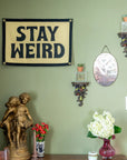 Stay Weird Camp Flag by Faire displayed on a green wall, adding playful style above floral and eclectic decor.