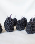 Eight Faire Blackberry Beeswax Candles stand upright on a soft, light surface. Each candle features a purple stem and is spaced unevenly in two loose rows with a white background.