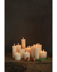 Creative Co-op Unscented Candle 4x4 candles in different heights are lit and placed on a rustic wooden surface, with two wilted red and yellow roses nearby against a dark background.