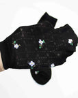 A hand displays a Faire Mini Floral Cotton Sock in black, featuring small white and yellow flowers; a matching sock is softly blurred in the background on a white surface.