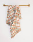 The Faire Cook's Cloth, a soft cotton-linen blend kitchen towel in muted green, orange, and beige plaid, hangs neatly over a brass towel bar on a plain white wall, combining style with durability.