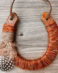 The Lucky Horseshoe by Faire is an upcycled keepsake wrapped in orange-brown yarn and twine, accented with a fluffy brown feather and a spotted feather, displayed on weathered wood.