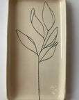 The Floral Small Tray by Faire is a rectangular cream ceramic piece featuring a minimalist hand-etched black line drawing of a leafy plant stem in the center, adding botanical style and detail.