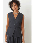 A woman with short curly hair wears the SUNCOO Derek Vest—a sleeveless, dark gray pinstripe piece woven with metallic thread—paired with matching pants and a beaded necklace, standing against a plain white background.