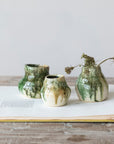 Three small Crackle Glaze Vases by Creative Co-op, in green and beige with a reactive crackle finish, sit on an open book atop wood. One holds dried flowers. The plain, neutral background highlights the organic shapes.