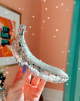 A hand with purple nail polish holds the Disco Banana by Faire—a banana-shaped decor piece covered in glass mirror tiles—casting shimmering reflections on a peach-colored wall in the background.