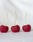 Faire’s Cherry Beeswax Candles feature three upright, cherry-shaped candles with green stems and a vivid red color, beautifully resembling real cherries on a white surface.