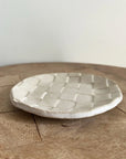The Faire Ghost Checker Trinket Dish, a small, round glazed stoneware dish with a woven checkerboard pattern, rests on a wooden surface against a plain, light-colored background.
