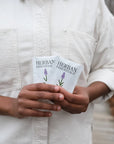 A person in a white shirt holds two packets of Faire Lavender Towelettes – 20 Individual Essential Oil Wipes, each with a lavender illustration, while standing outdoors on a wooden path amid autumn foliage.