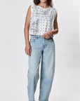 A woman wears the Faire Hemp Jackie Pleat Shell Bandana, a sleeveless white hand-printed shirt with blue paisley and star patterns, paired with light blue high-waisted relaxed-fit jeans, standing against a plain white background.