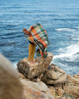 A person stands barefoot on a rocky ocean cliff, wrapped in the Faire Vrou-Vrou blanket with bold textile design blowing in the wind, as vivid blue waves and swirling seaweed move below.