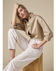 A woman with blonde hair sits on a wooden block against a beige background, wearing The Everything Shirt by Sold Out NYC—an oversized 100% cotton button-up—paired with white pants. She gazes at the camera, resting her elbow on her knee and hand on her head.