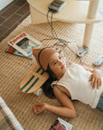 A girl in a beanie and tank top relaxes on a woven rug with headphones, resting her head on the BANWOOD SKATEBOARD - GREEN by Faire. Vintage video game controllers, a cassette player, and skateboard magazines are nearby.