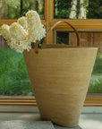 An Indaba Hydrangea Paniculata Stem is arranged in a large woven basket with a leather handle, set indoors near a window overlooking sunlit green trees.