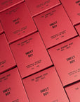 Several red boxes arranged in a grid display "SWEET BOY" and "THE SENSORY RIOT EST. 2017." Faire’s SWEET BOY 12 oz candle is a clean-burning, non-toxic scented soy wax candle designed to be intentionally unexpected (340g).